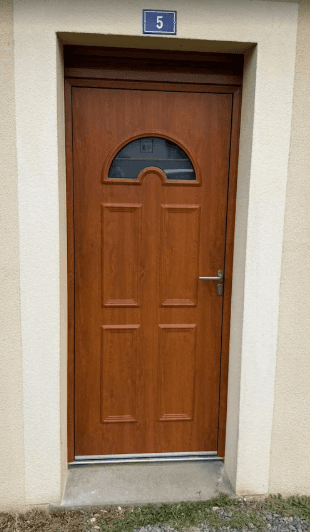 Porte d'entrée marron effet bois, fenêtre en demi-lune, quatre panneaux et numéro cinq au-dessus.