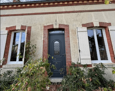 Façade beige avec porte grise, briques rouges et fenêtres blanches aux volets blancs ouverts.