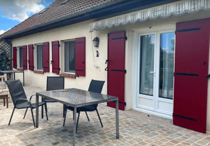 Terrasse pavée avec table et chaises devant une maison blanche aux volets rouge bordeaux.