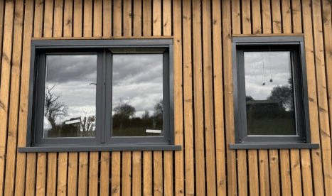 Mur en bois à lattes verticales avec deux fenêtres grises reflétant un ciel nuageux.