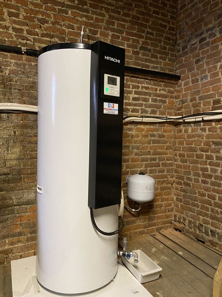 Grand chauffe-eau Hitachi blanc installé dans une pièce avec des murs en briques apparentes.