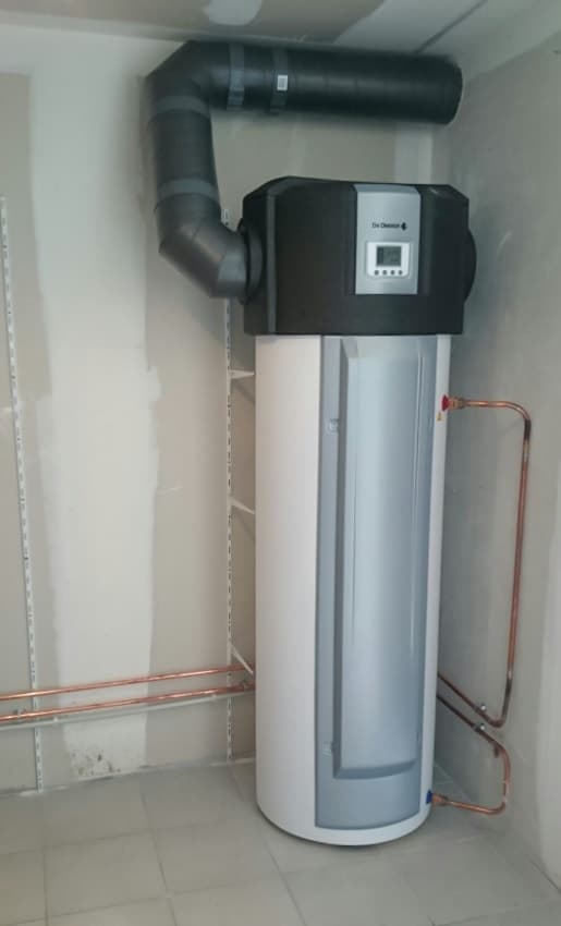 Chauffe-eau thermodynamique De Dietrich blanc avec conduit noir et tuyauterie en cuivre apparente.