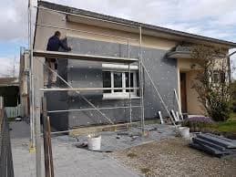 Ouvrier installant des panneaux d'isolation gris sur la façade d'une maison depuis un échafaudage.