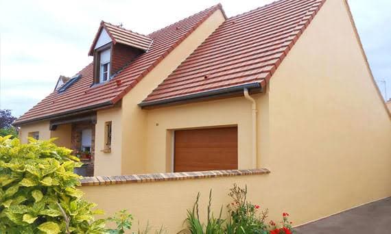 Maison moderne à façade beige, toit en tuiles rouges, garage et arbustes au premier plan.