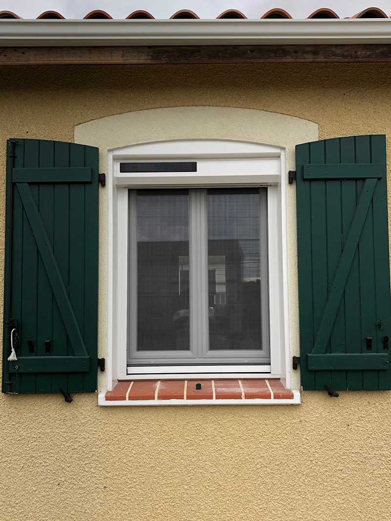 Fenêtre à cadre blanc, volets verts ouverts et volet roulant solaire sur façade jaune.